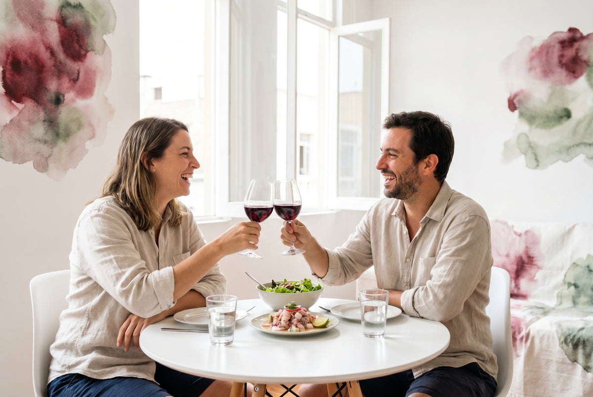 Pareja de unos 35 años brindando con copas de vino tinto seco en una mesa luminosa con cebiche y ensalada fresca