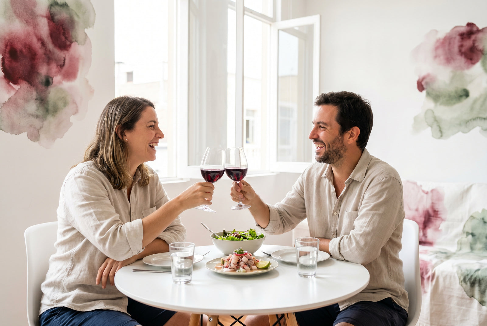 Pareja de unos 35 años brindando con copas de vino tinto seco en una mesa luminosa con cebiche y ensalada fresca