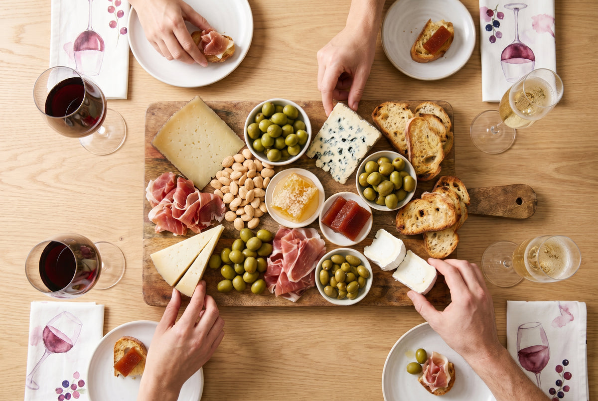 Tabla de quesos y tapas con copas de vino tinto y Cava, jamón, aceitunas y pan, en una mesa luminosa lista para un picoteo entre amigos